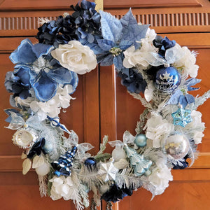 Large grapevine wreath covered with flocked pine sprigs. Navy, powder blue, aqua, silver and white ornaments decorate the wreath. The ornaments are tucked in amongst a collection of flowers including,  blue velvety magnolia and poinsettia blooms with dark navy , cornflower blue and white hydrangeas. Glittery silver feathers swirl around the ornaments in this elegant , floral Christmas wreath