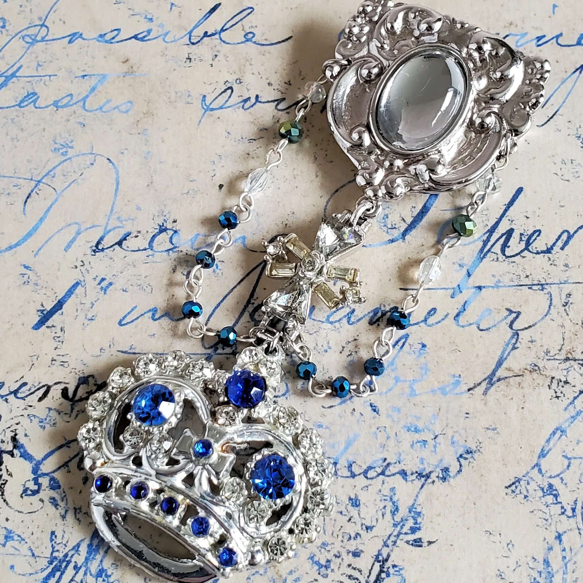 Three tier brooch pin with a blue and clear rhinestone crown. The crown is topper with a vintage crystal Maltese cross pendant and finished with a Victorian style pendant with a crystal stone centerpiece. a blue and crystal bead chain accents the piece.