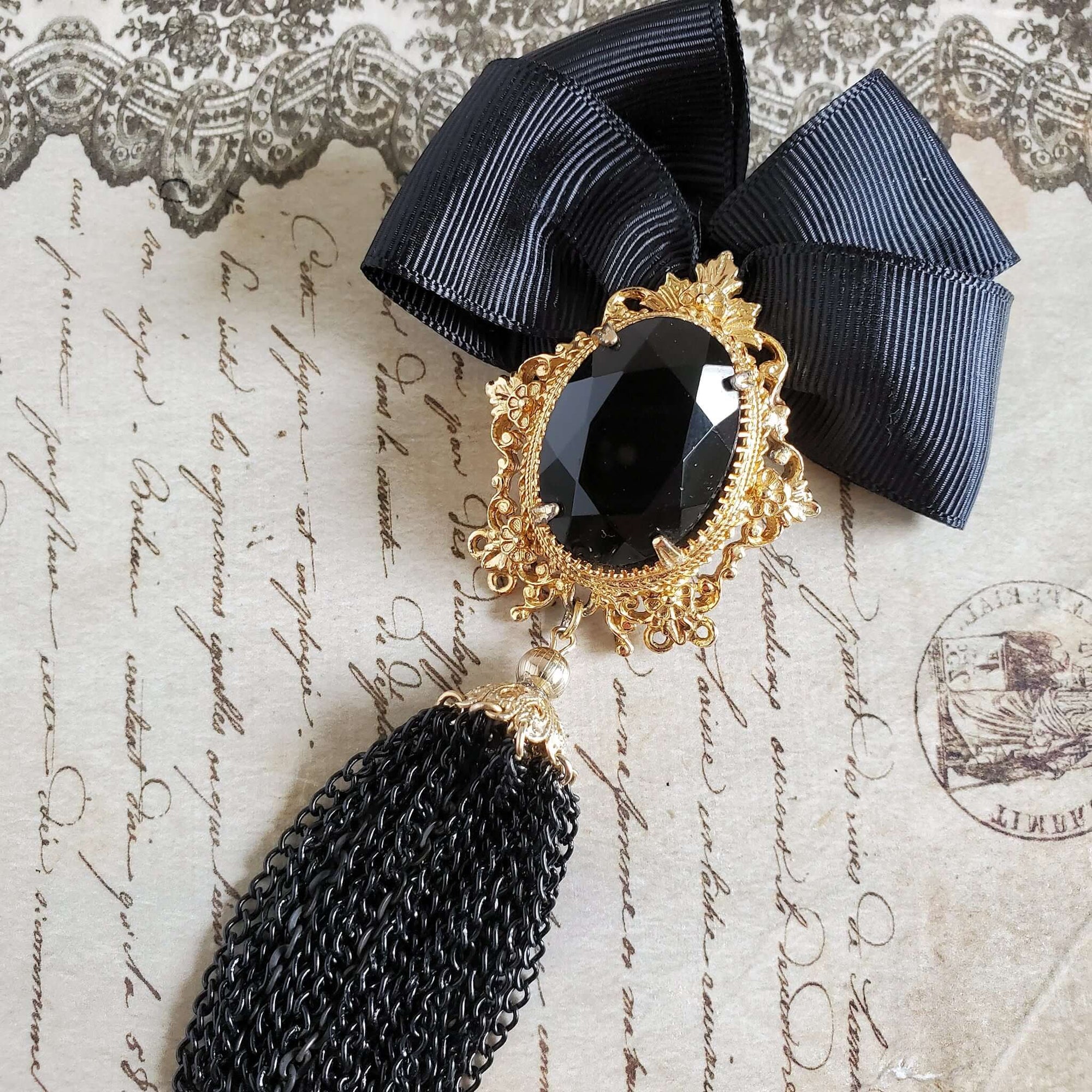 Designer brooch with black ribbon bow and a large stone set in a gold filigree setting. The brooch is finished with a black chain tassel topped with a gold bead cap.