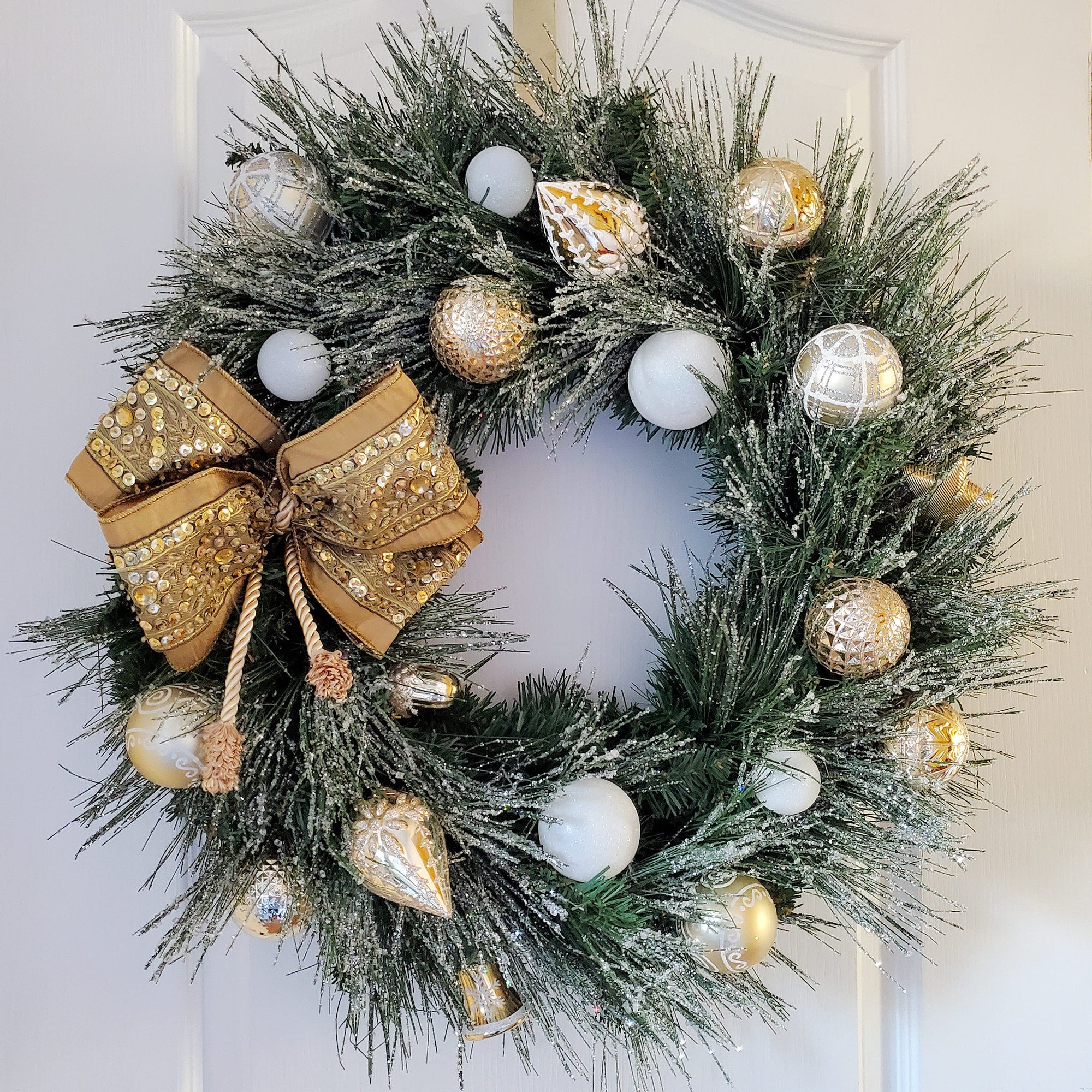Iced Pine Branches Circle the Wreath. The wreath is decorated with a collection of gold and white sparkling ornaments.
