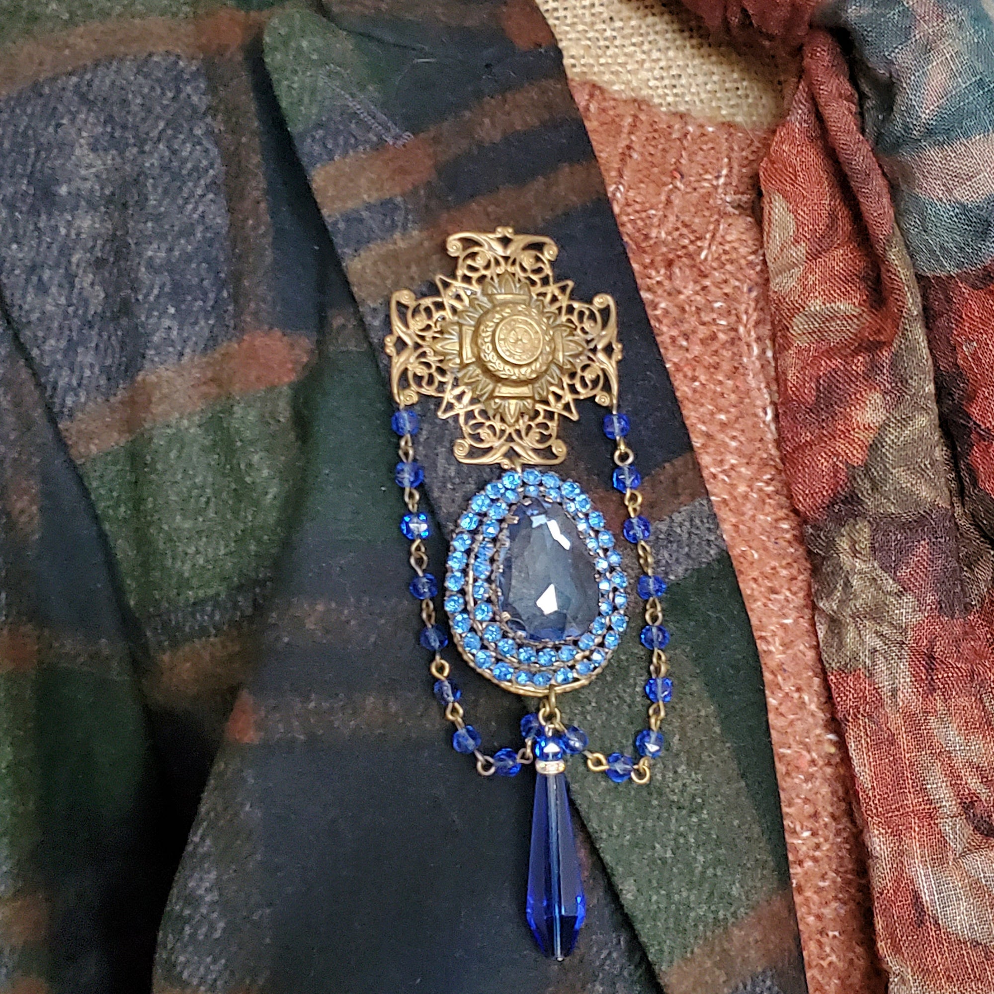 Victorian Style Brooch with an Antique Brass Filigree Maltese Cross, with a genuine military pin center. A vintage large blue crystal dress clip is the statement centerpiece. Two strands of antique blue glass beads and a 1920s faceted teardrop blue bead finish this brooch.